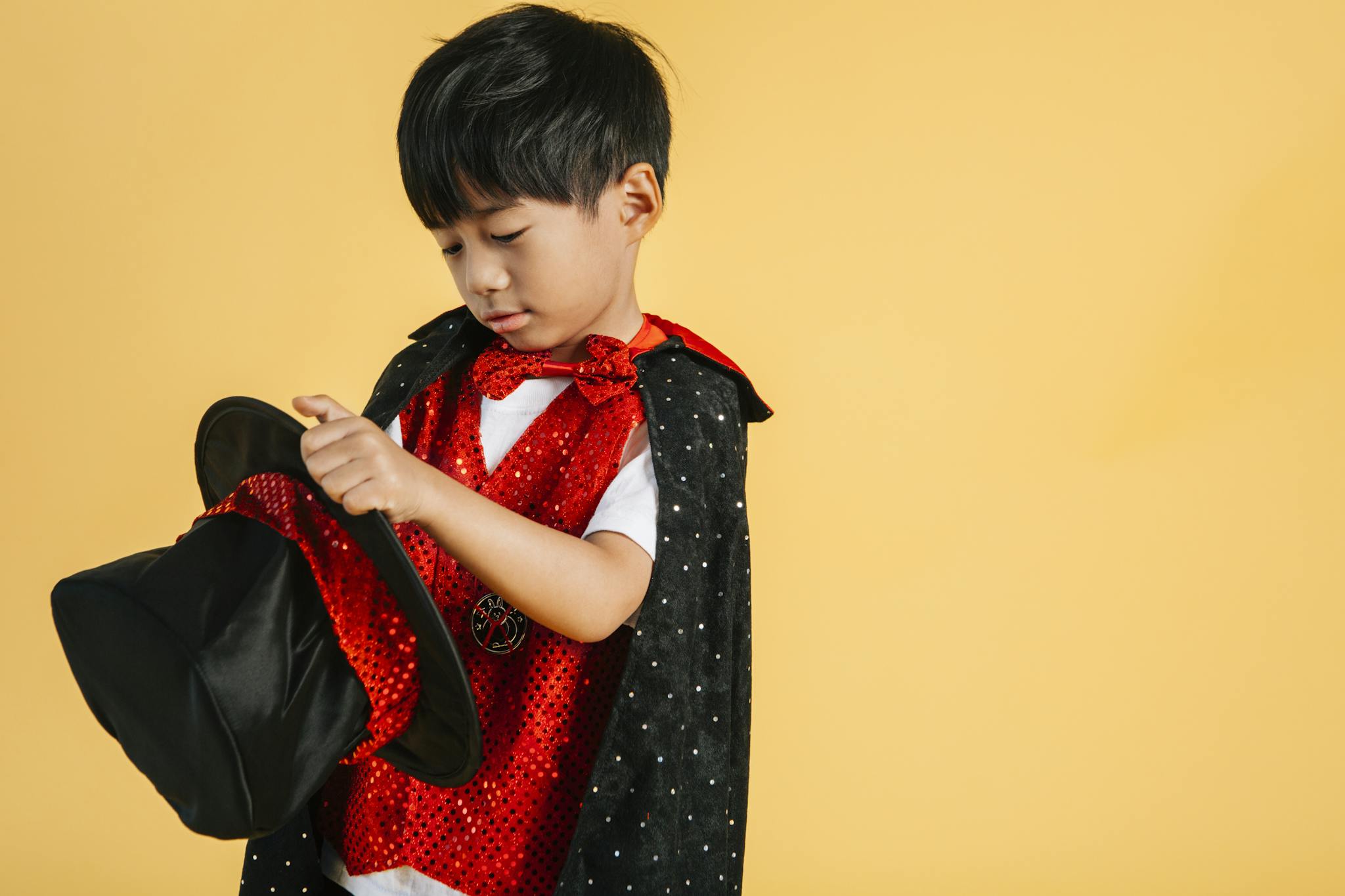 Child dressed as a magician in a sparkly costume holding a hat against a yellow background.